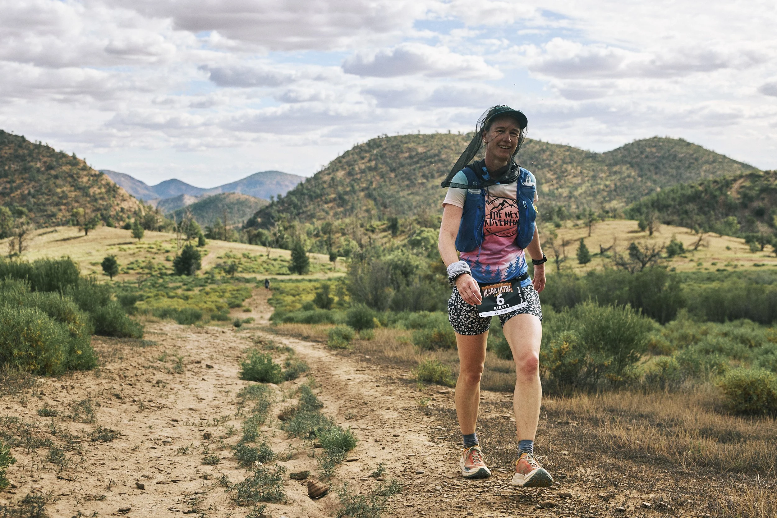 A woman smiling and running on a dirt trail in a mountainous area with green shrubbery and trees, wearing athletic clothing and a hydration pack.