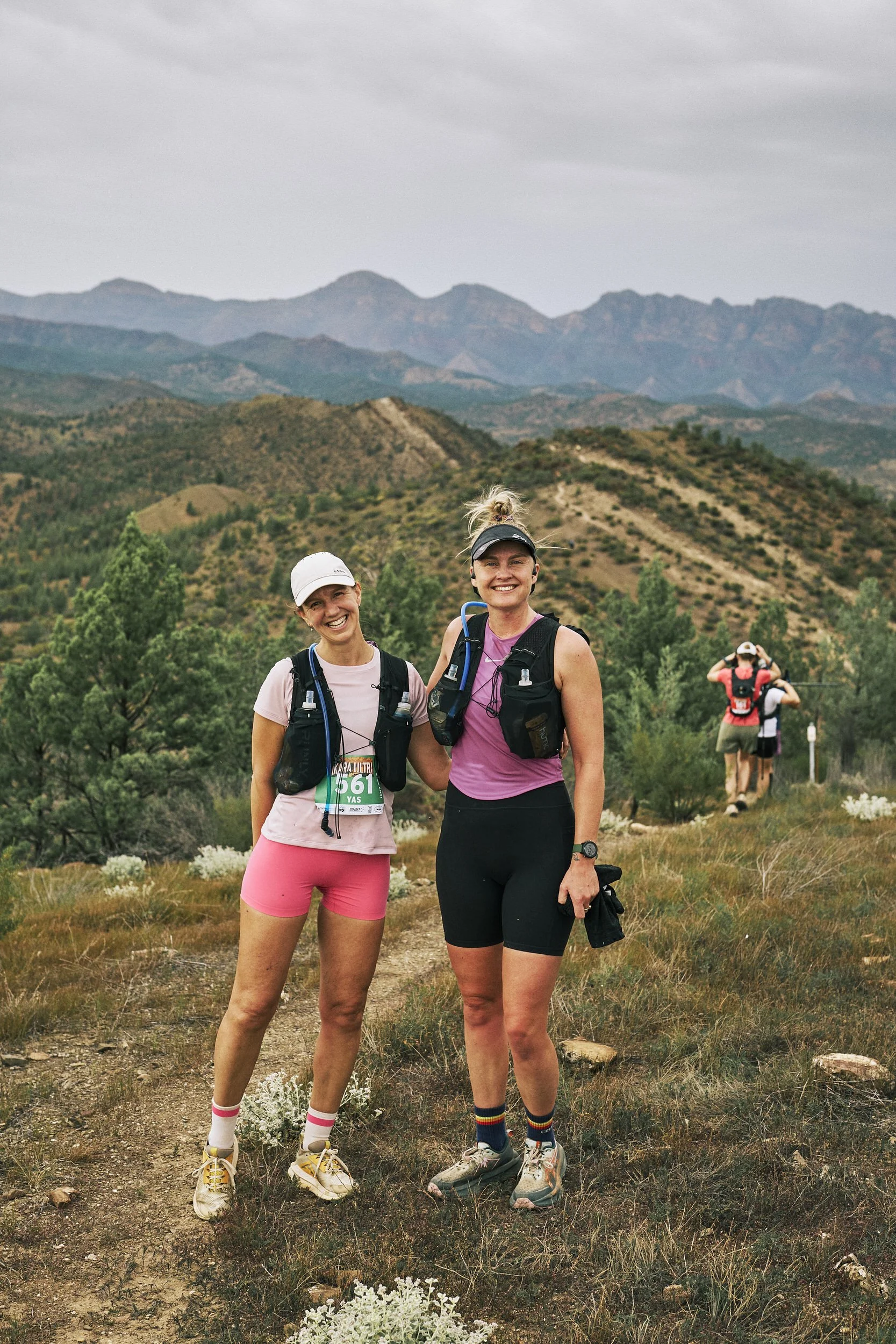 Two women trail running on a dirt path in a desert landscape with mountains in the background, wearing outdoor athletic gear.