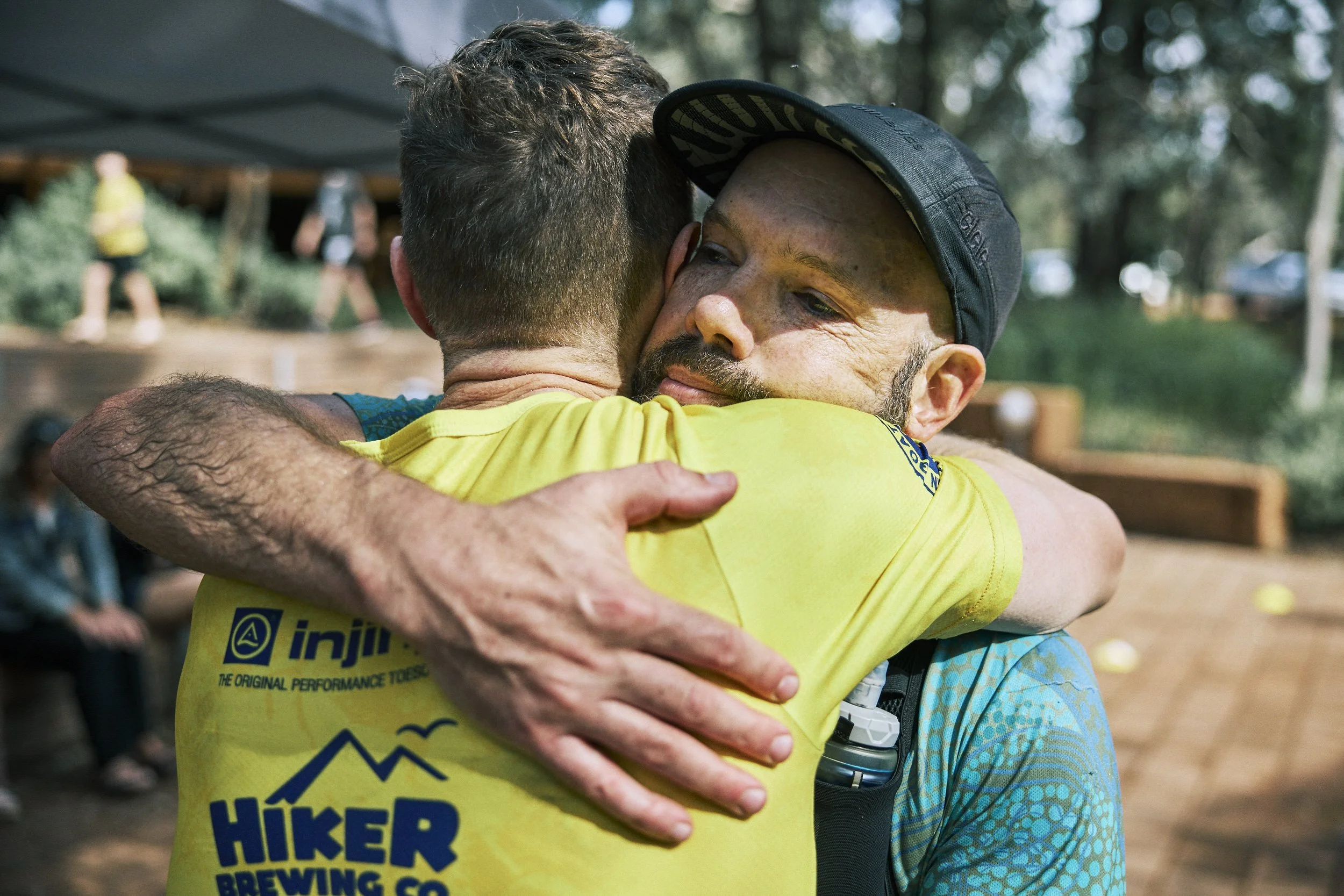 Two men hugging each other outdoors, one with a cap and a yellow shirt, the other with a short beard and a blue patterned shirt.