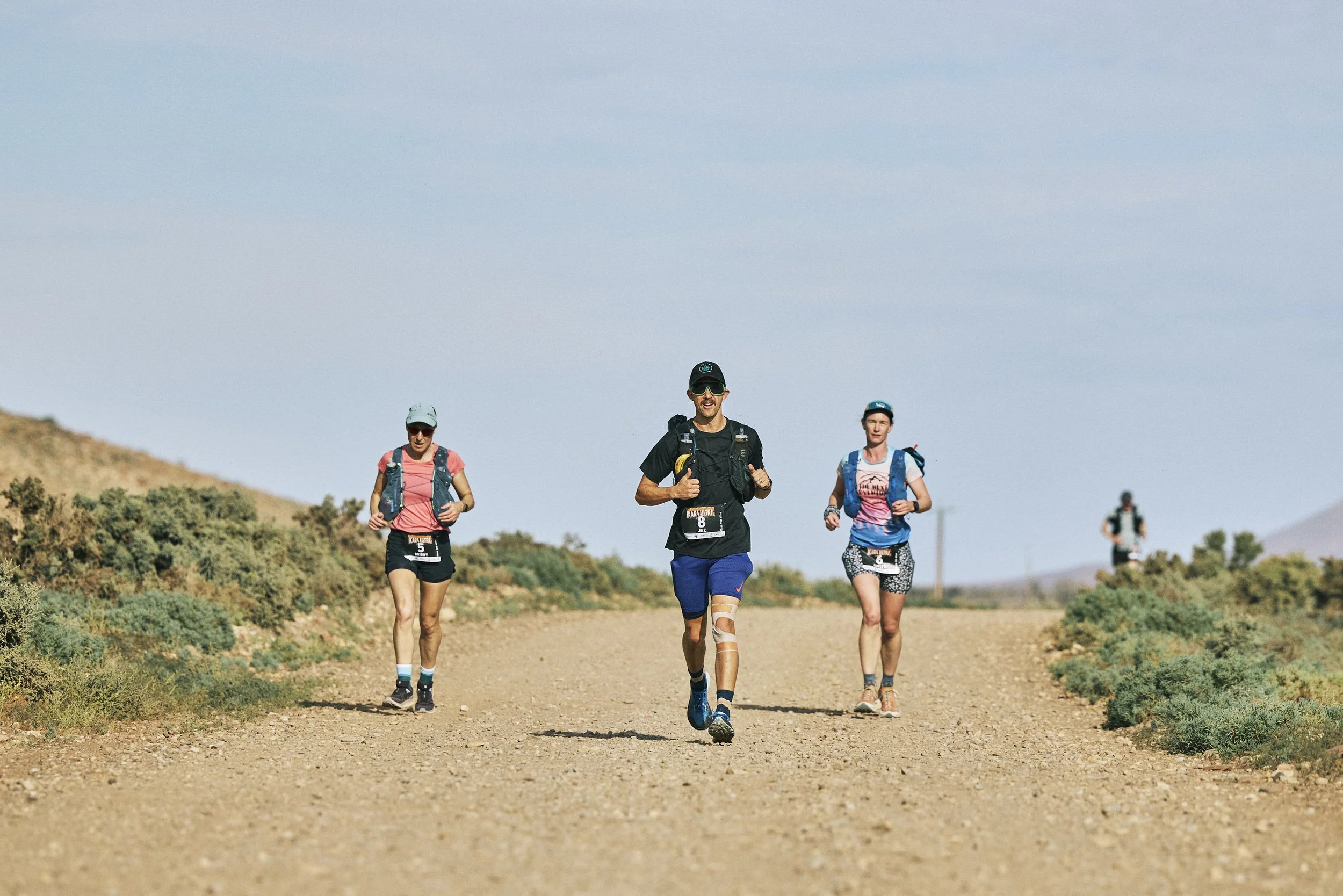 Group of four runners participating in a trail race in a desert landscape with low bushes, blue sky, and distant hills.