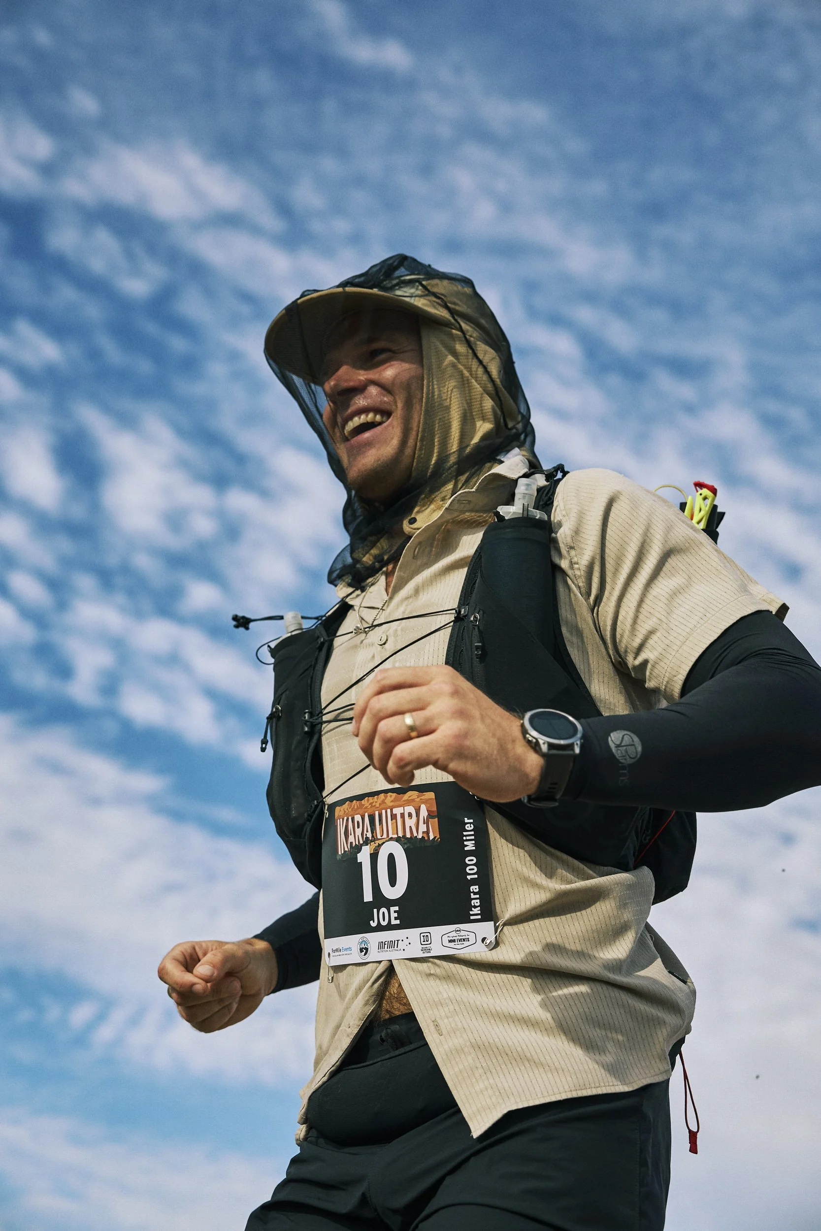 A man running outdoors with a race bib that reads 'JOE' and the number 10, wearing a hat, long sleeve black shirt, and a beige vest with a hydration pack, smiling with a cloudy sky in the background.