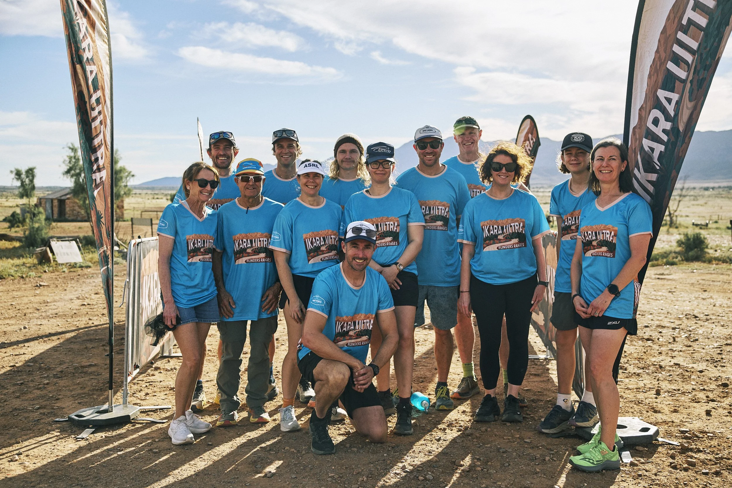 Group of people in athletic clothing standing and kneeling outdoors, posing for a photo at a race event with flags and a rural landscape in the background.