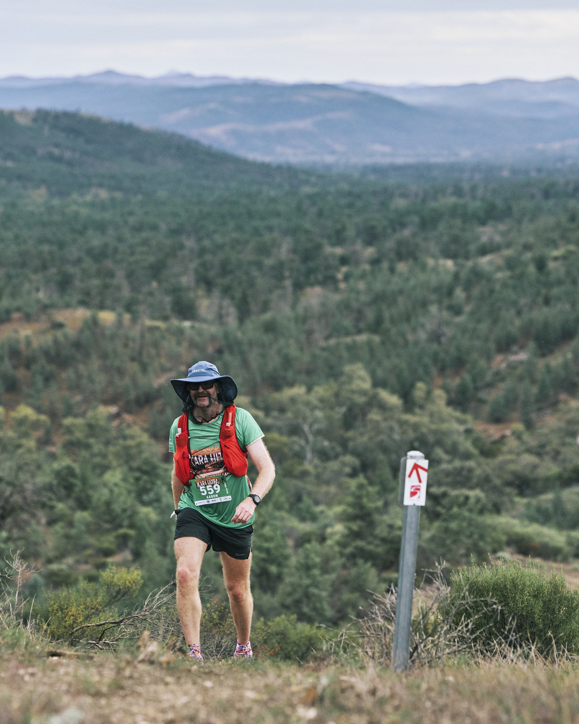 A male trail runner with a race bib, wearing a wide-brimmed hat and sunglasses, running along a mountain trail with hills and a signpost in the background.