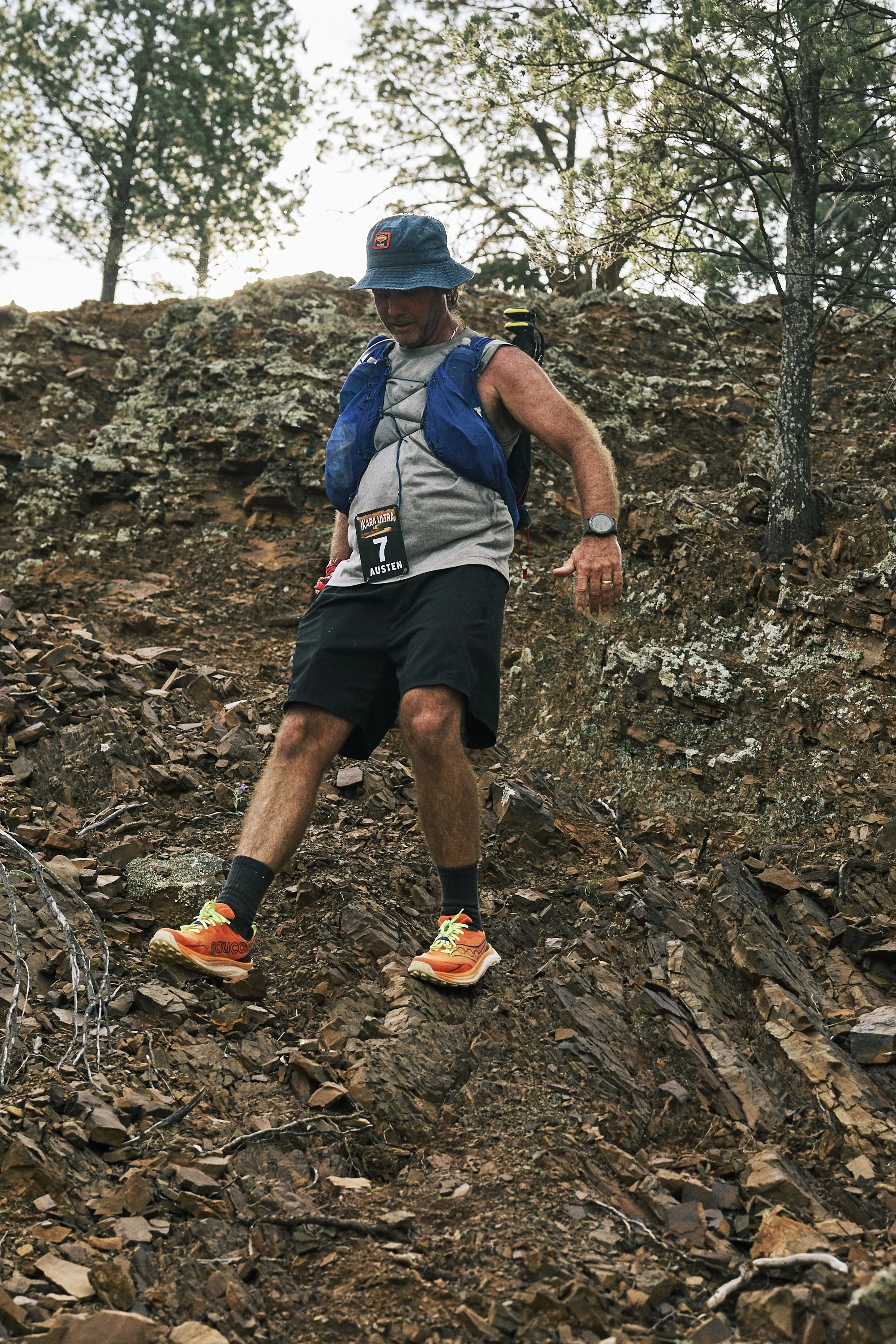 A man in athletic gear, wearing a blue hat and a race bib numbered 7, balancing on rocks on a sloped outdoor trail surrounded by trees.