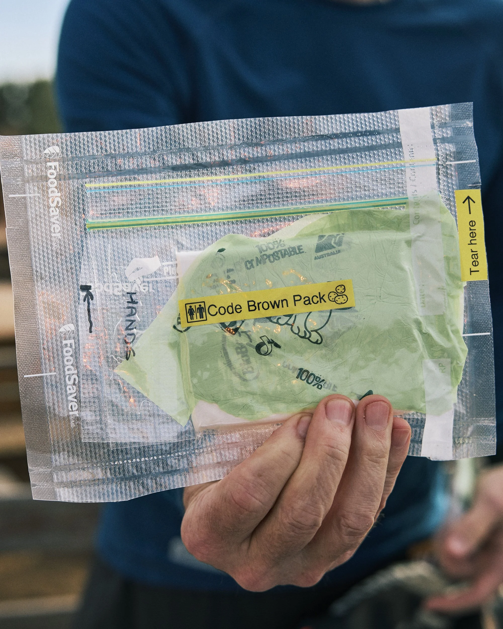 A person's hand holding a clear plastic package containing a yellow banana with a green sticker that reads '100% biodegradable.' The package has a label with a yellow background and black text that says 'Code Brown Pack' with a small icon of a person