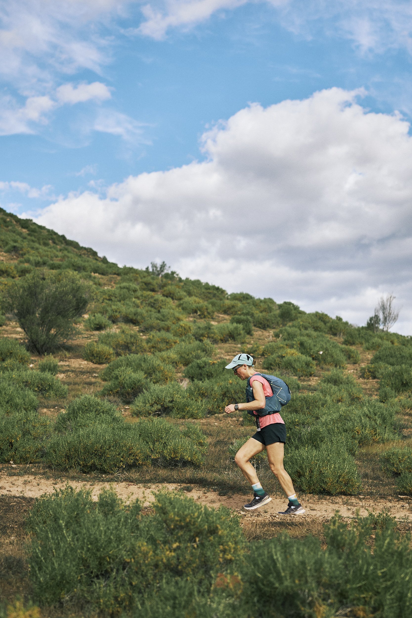 A woman trail running in a green, hilly landscape under a partly cloudy sky, with a blue and gray cap, pink shirt, black shorts, and a small backpack.