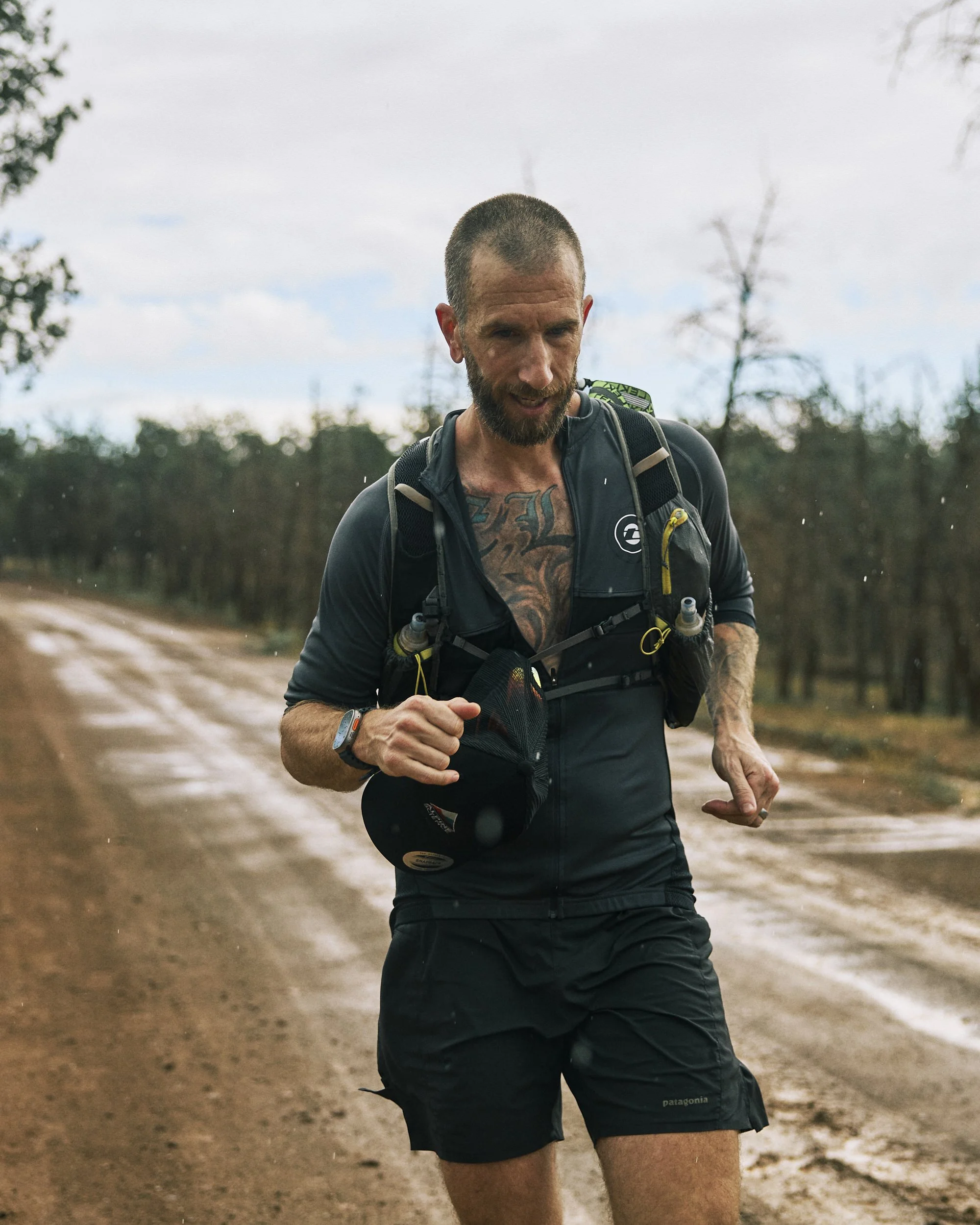 A man running outdoors during the daytime, wearing a black athletic top, black shorts, and a hydration backpack. He has a short haircut, a beard, and tattoos on his chest visible through his shirt. The background features a dirt trail with trees and 
