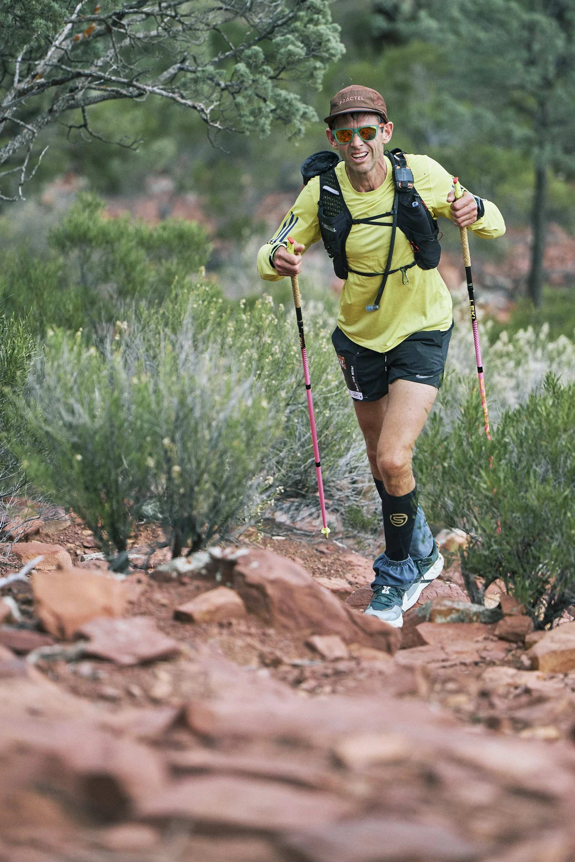 A man trail running on a rocky dirt path through a green forest, wearing a yellow shirt, black shorts, blue shoes, sunglasses, a brown hat, and carrying a backpack with hiking poles.