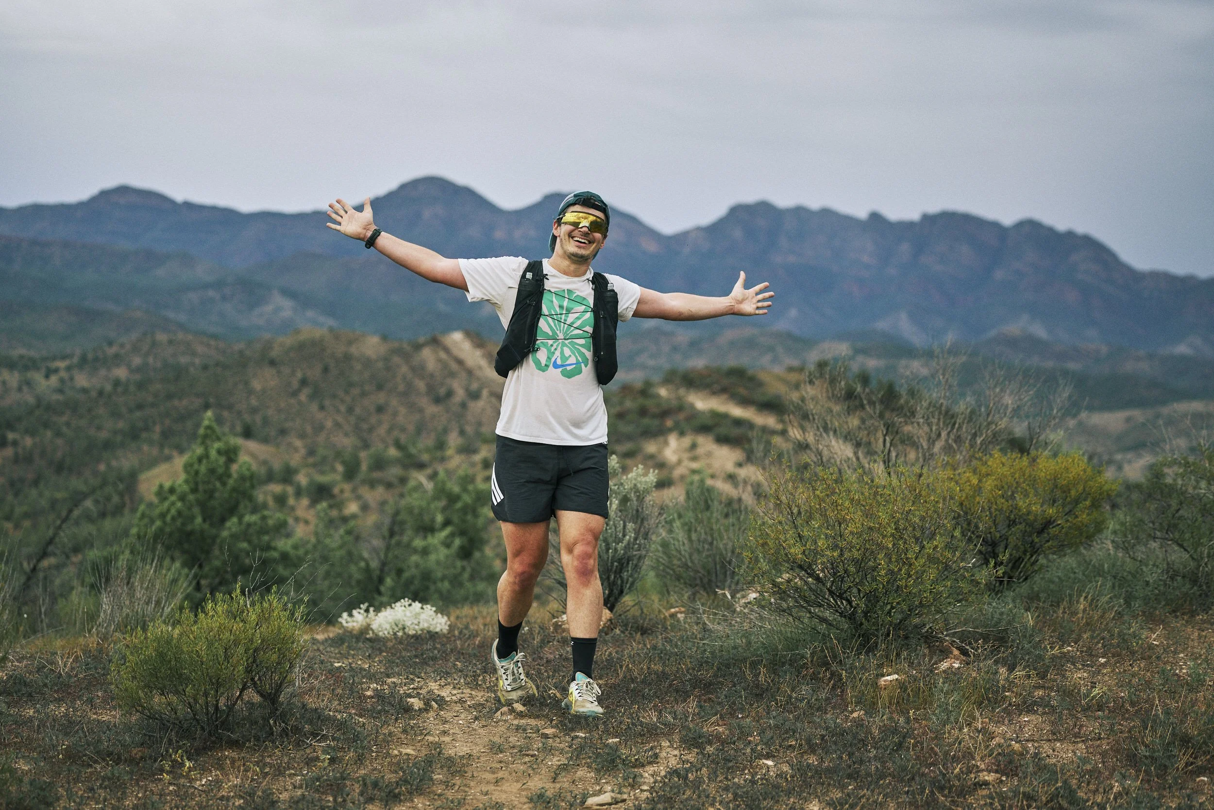 A man in athletic gear smiles with arms outstretched while hiking on a trail in a mountainous area surrounded by shrubs and low vegetation.