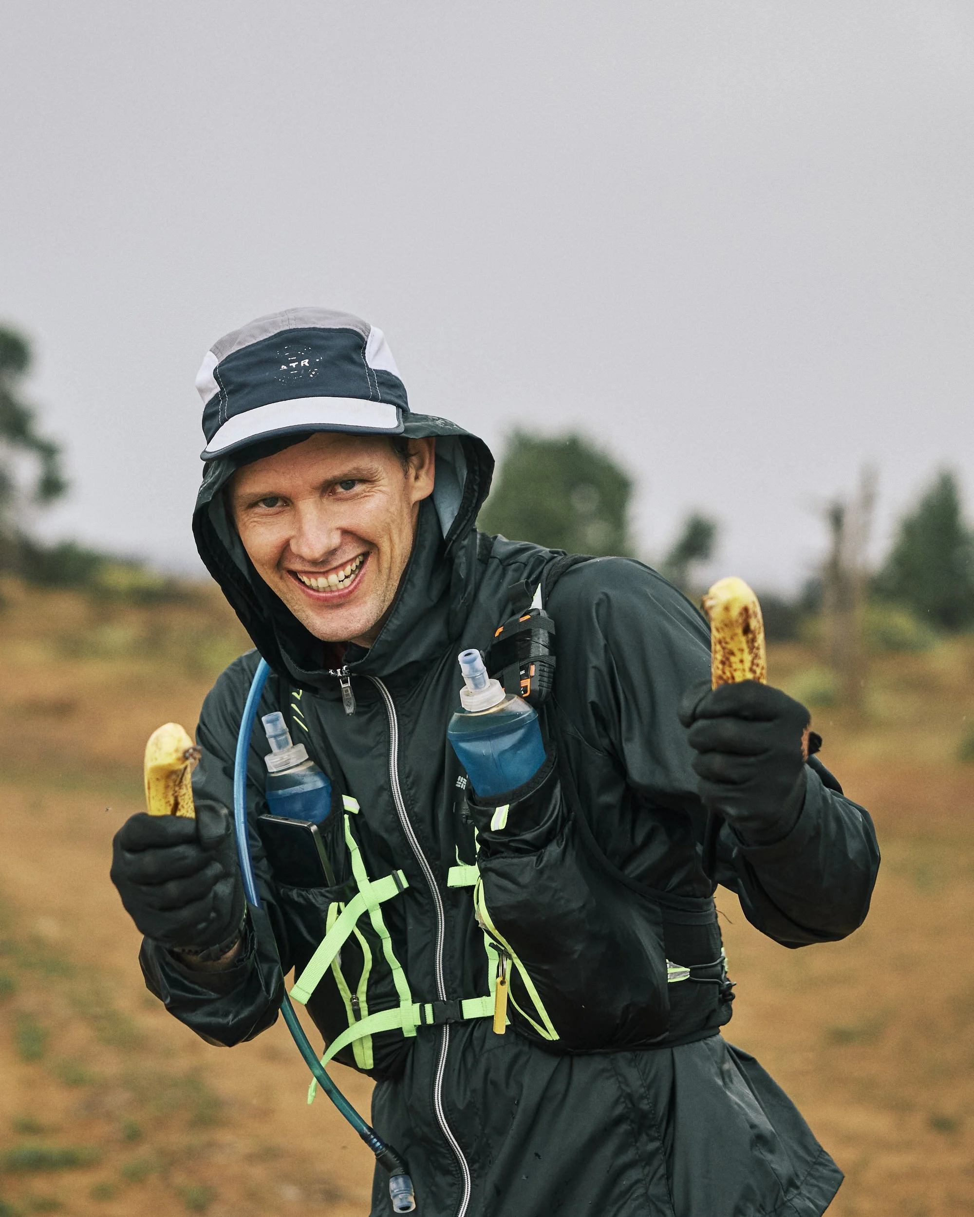A man running outdoors on a cloudy day, wearing a black rain jacket, a black and white hat, and holding a banana in one hand and a water bottle in the other.