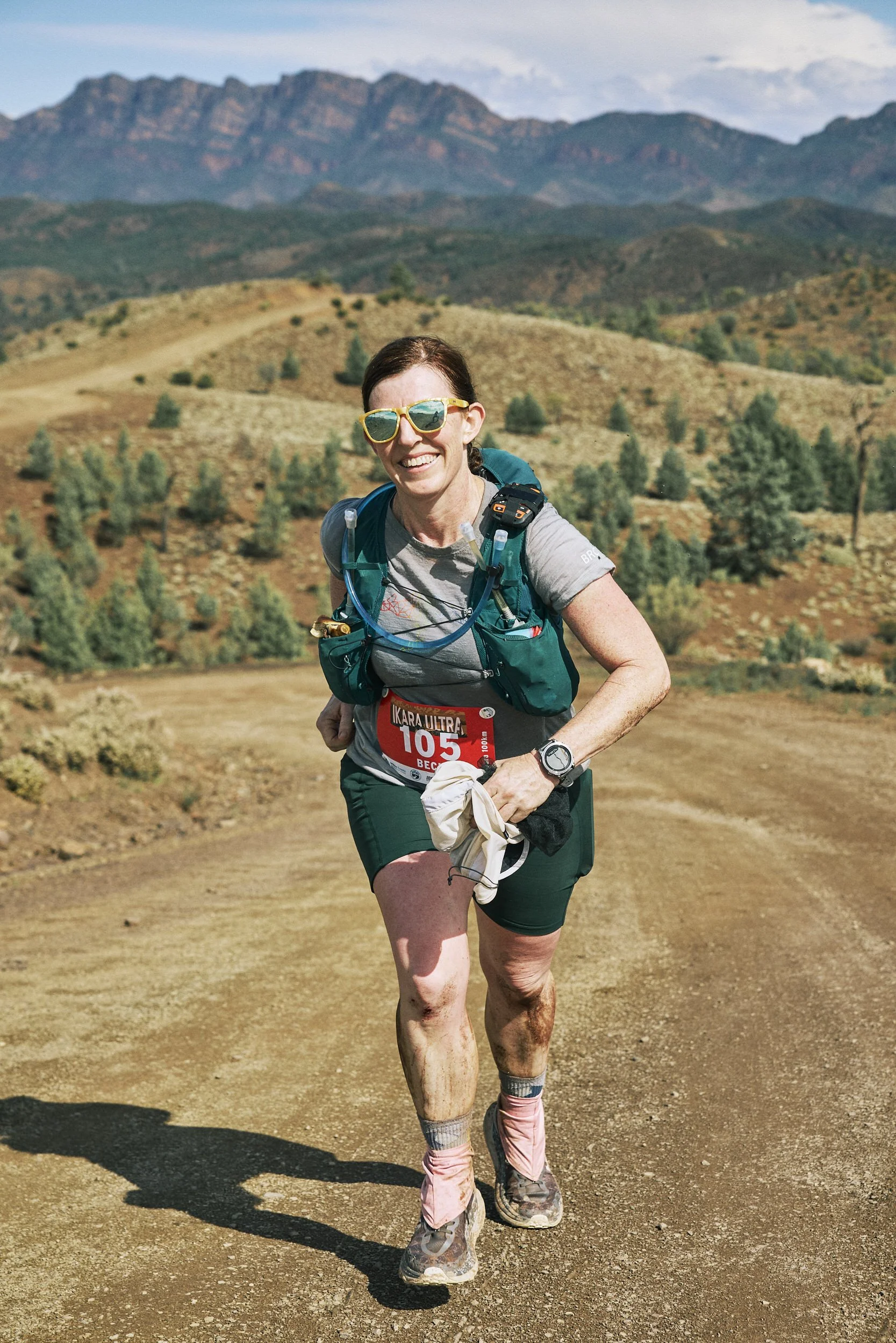 A woman smiling while running outdoors on a dirt trail in a mountain landscape, wearing sunglasses, a gray t-shirt, green shorts, a hydration pack, and running shoes.