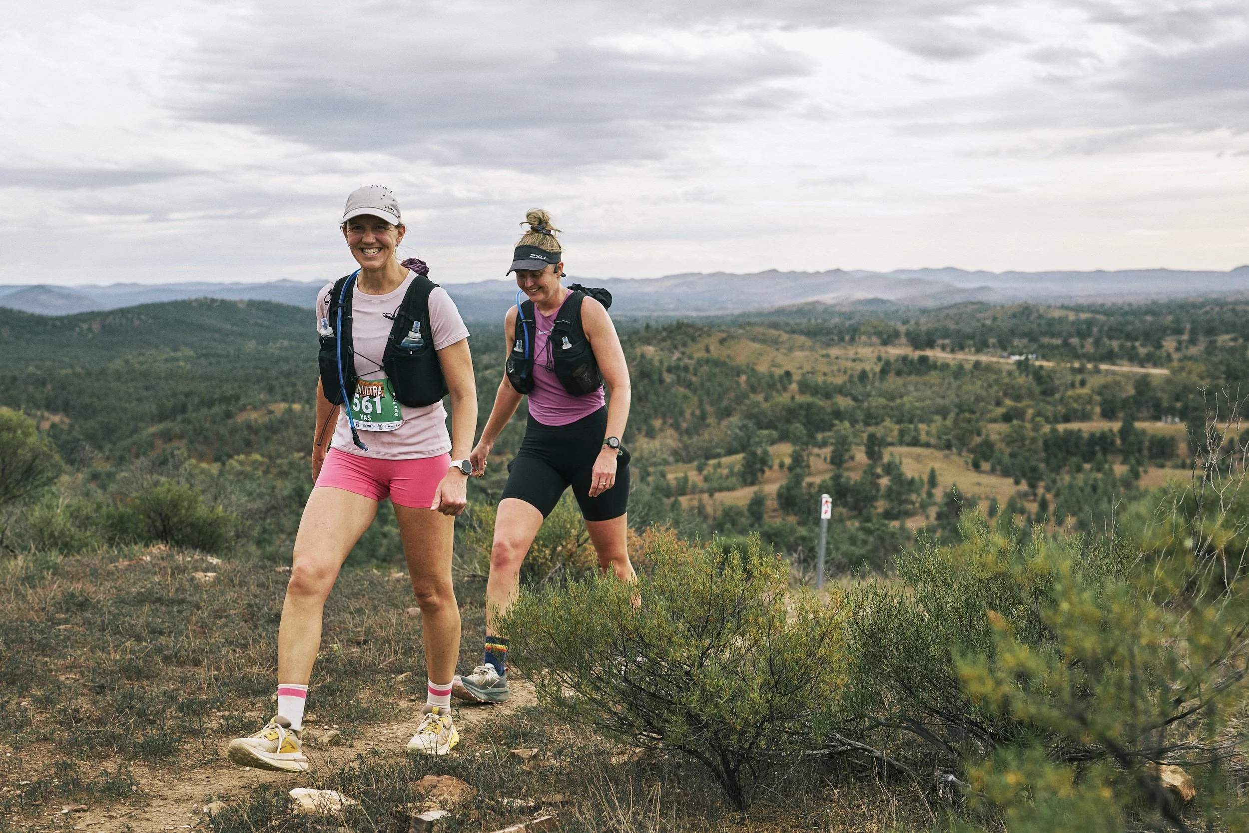 Two women trail running on a dirt path in a mountainous landscape with cloudy sky.