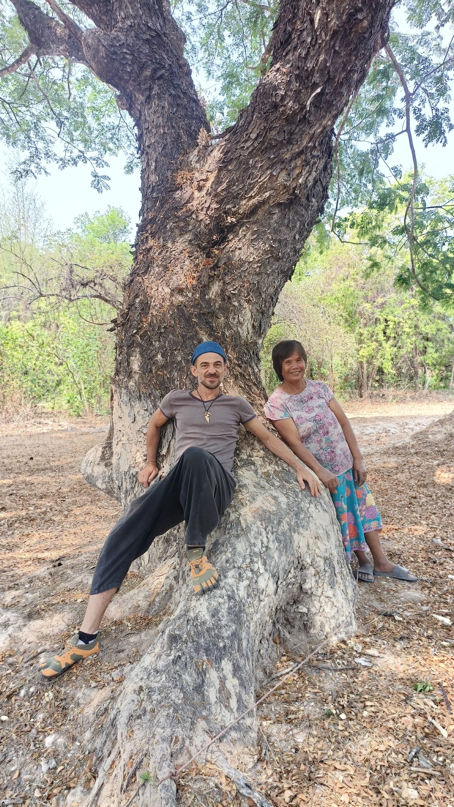 Deux personnes, un homme et une femme, posent devant un grand arbre dans une forêt, assis sur une racine d'arbre.