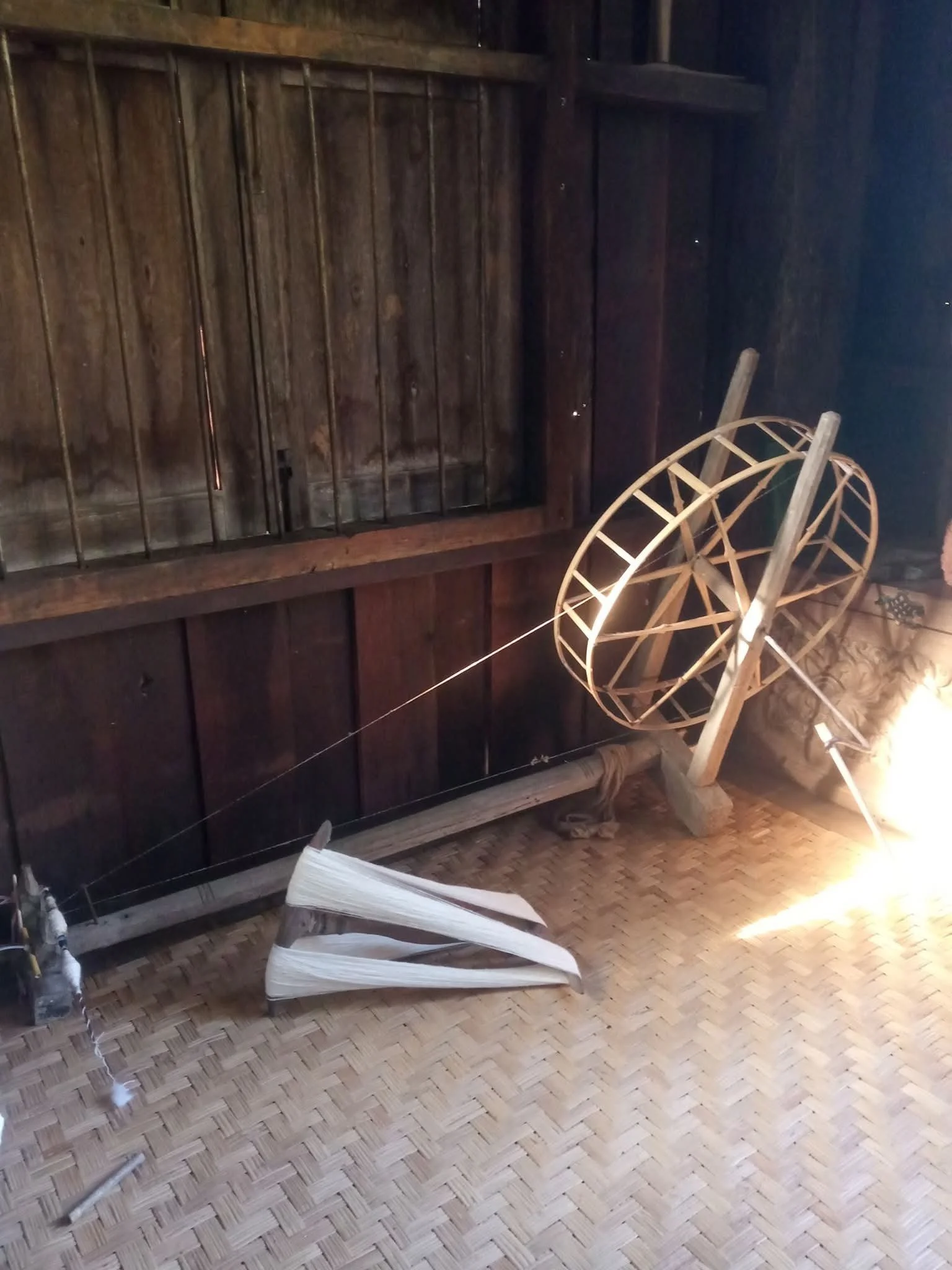 Une structure en bois en forme de roue ou d'hélice, posée à côté d'un objet en bois, dans une pièce avec un mur en bois sombre et un sol en tuiles.