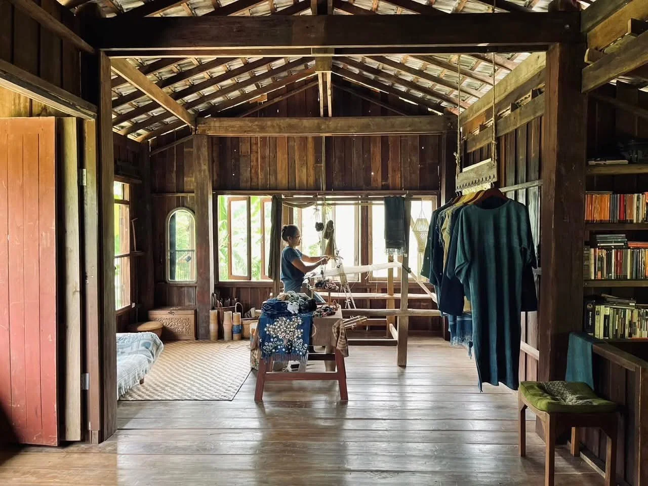 Une femme fait de la teinture ou du tissage dans une pièce en bois avec de grandes fenêtres, vêtements suspendus, étagères avec des livres, un petit lit, et un tapis beige, dans un intérieur rustique et lumineux.