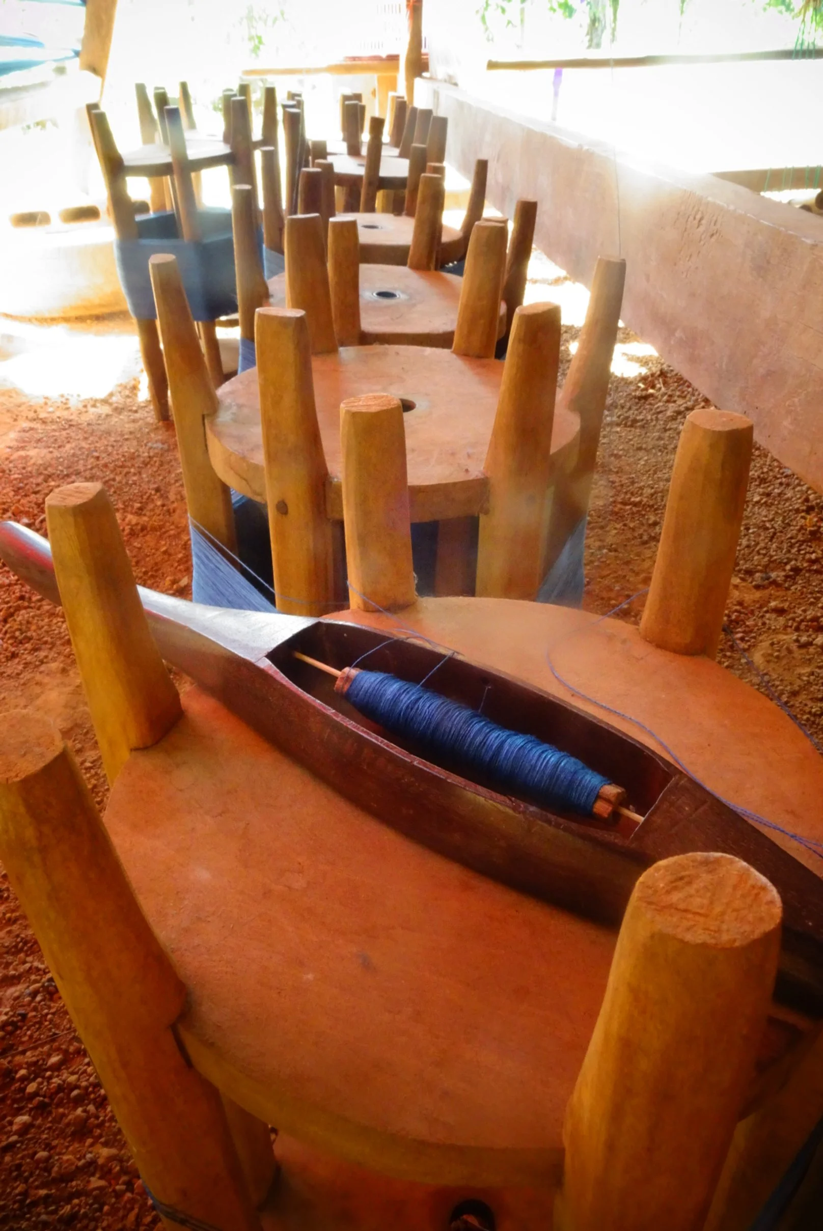 Une rangée de chaises en bois retournées et une navette de tissage en bois contenant une bobine de fil bleu indigo, dans un espace éclairé en plein air.