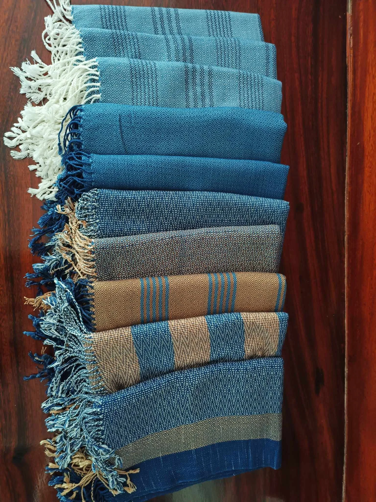 Une collection de huit étoffes ou tapisseries en tissu, aux motifs variés en nuances de bleu, beige et marron, avec des franges aux extrémités, disposées côte à côte sur un sol en bois.