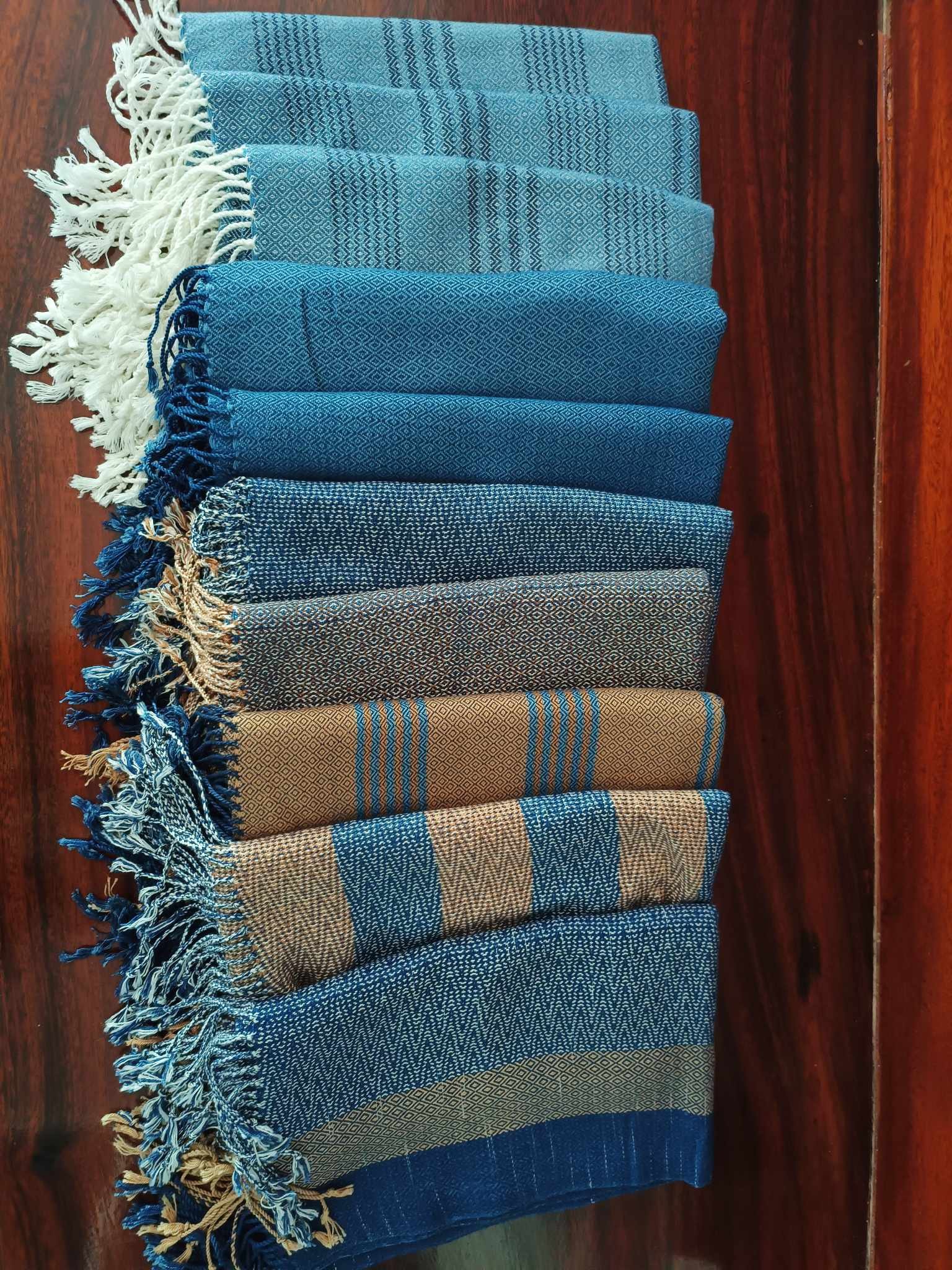 Une collection de huit étoffes ou tapisseries en tissu, aux motifs variés en nuances de bleu, beige et marron, avec des franges aux extrémités, disposées côte à côte sur un sol en bois.