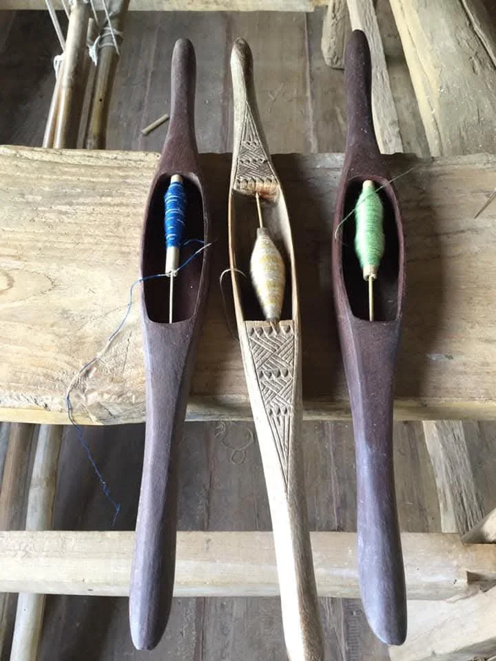 Trois outils en bois longs contenant des bobines de fil à tisser, posés sur une surface en bois.