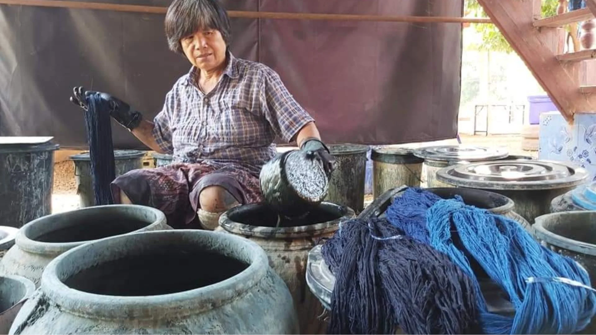Personne assise avec des seaux et des pots de terre cuite, en train de teindre des tissus en crème de colorant noir, dans un environnement intérieur ou extérieur.