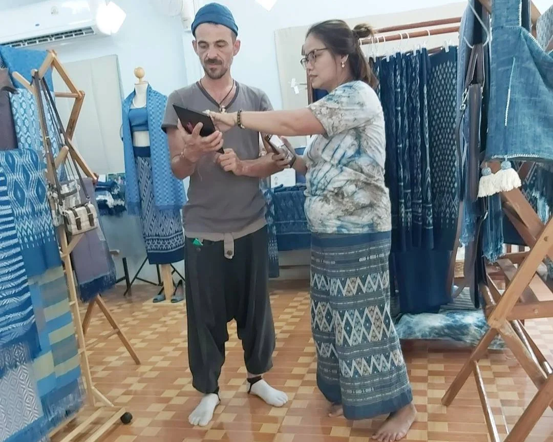Deux personnes discutent dans un magasin de textiles, entourées de tissus en différentes nuances de bleu exposés sur des étagères et des chevalets.
