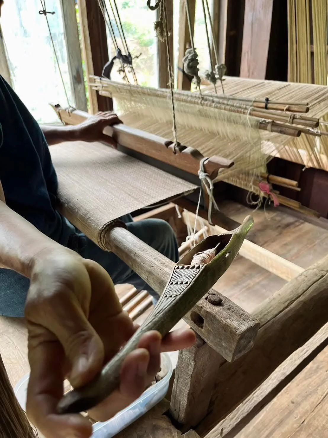 Une personne utilise un bâton tissé en bois pour travailler avec un métier à tisser traditionnel en bois, dans une pièce en bois avec lumière naturelle.