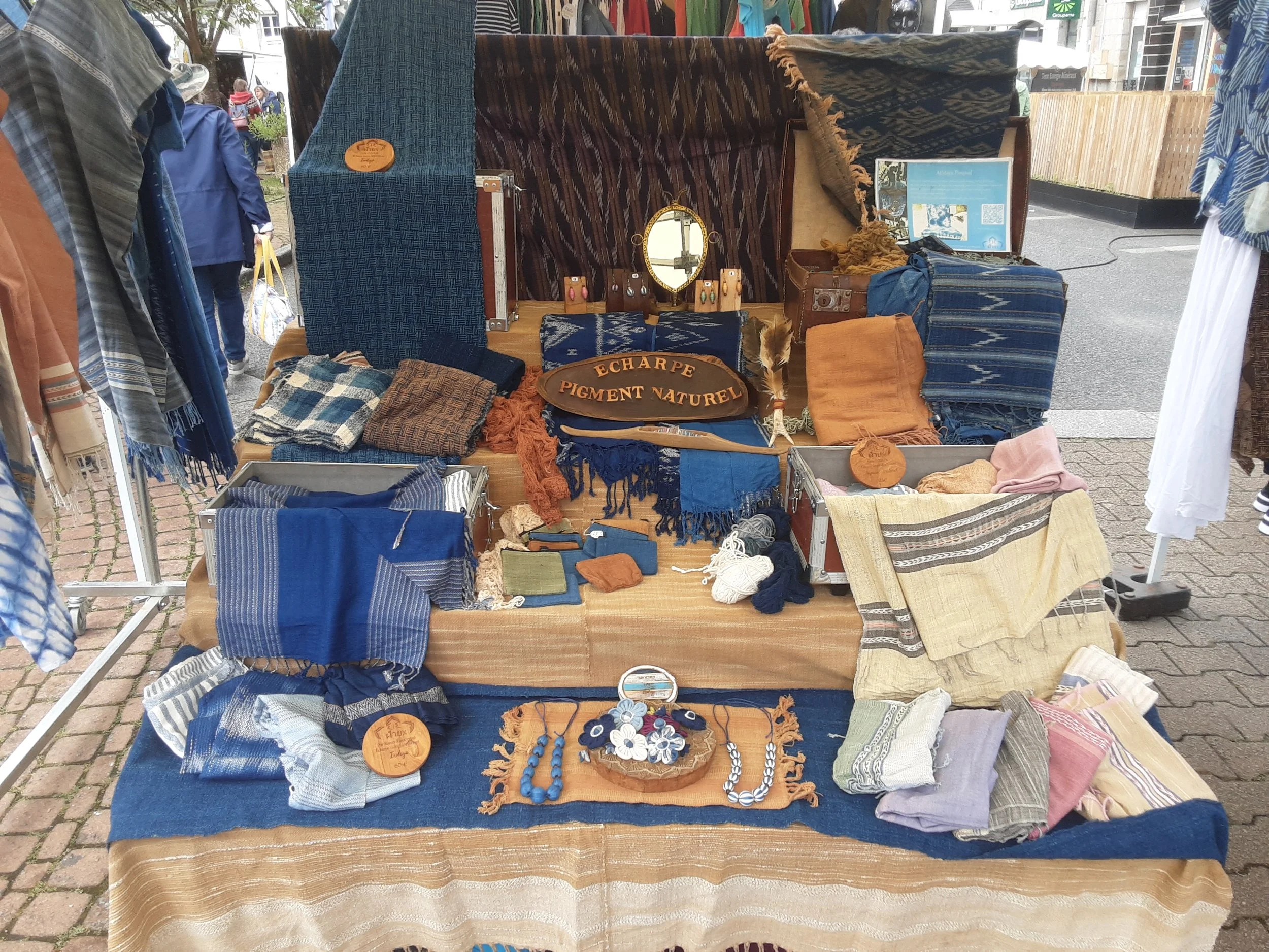 Vitrine de broderies, écharpes et accessoires en tissus en rapport avec les teintures naturelles, avec des objets en bois et des bijoux artisanaux.