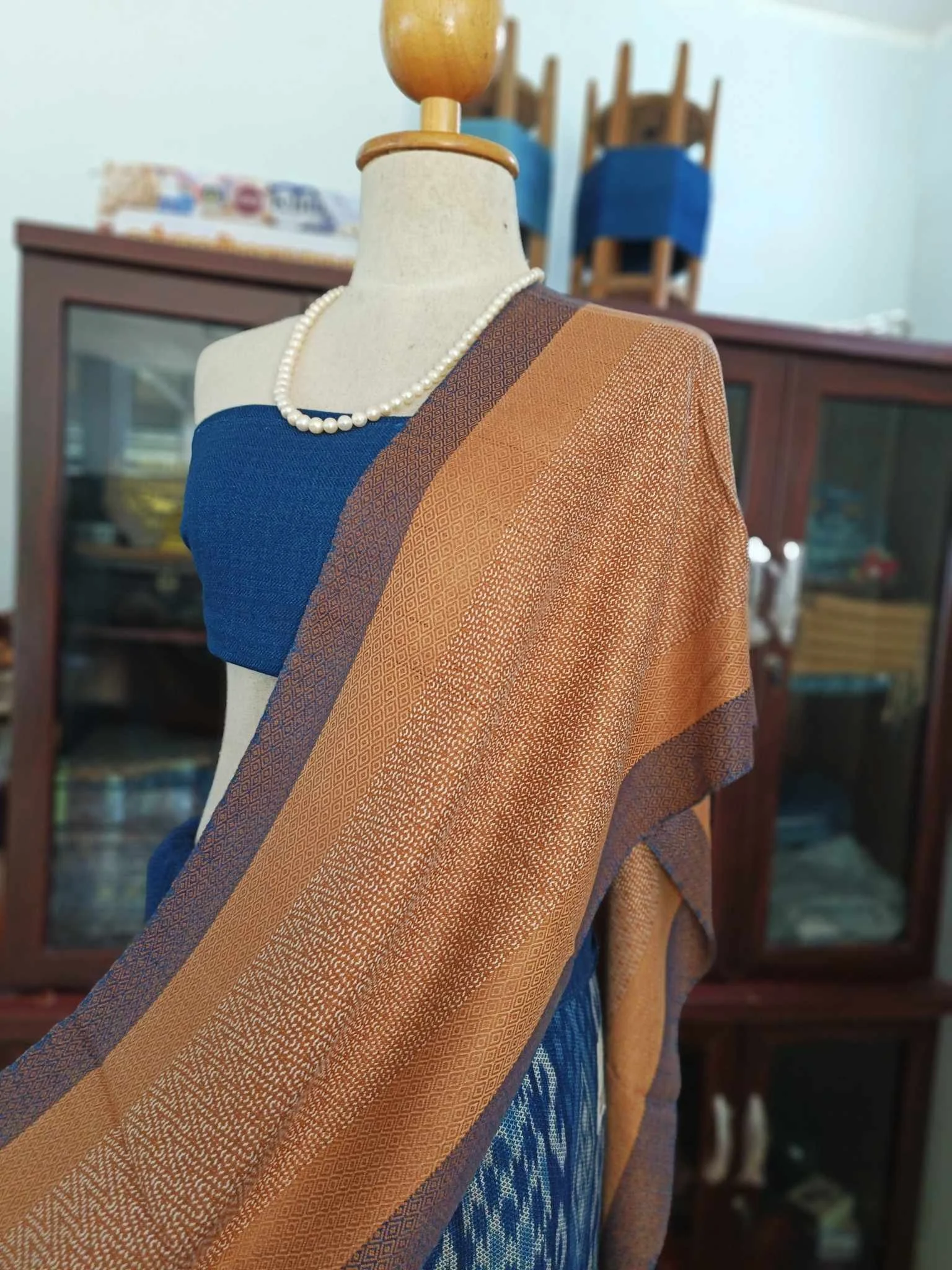 Mannequin portant un vêtement en tissu bleu et une écharpe en tissu orange avec motifs, porté avec un collier de perles.