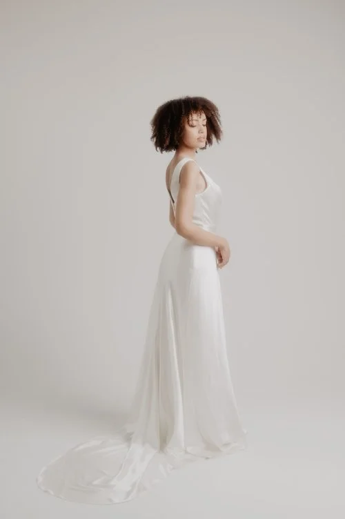 Sarah Barker minimalist satin A-line wedding dress with a plunging back and elegant long train, photographed in a soft neutral studio setting for a modern bridal look.