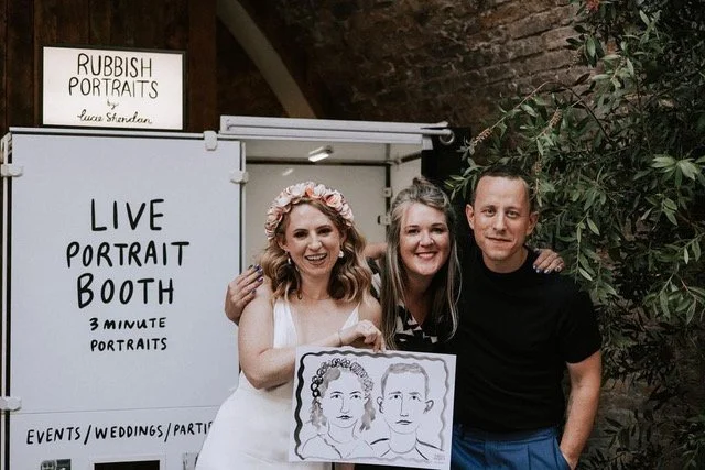 A couple and their guest pose proudly with their quirky hand-painted portraits outside the Rubbish Portraits live portrait booth by Lucie Sheridan. This fun and creative wedding entertainment gives guests a unique keepsake to take home while adding a