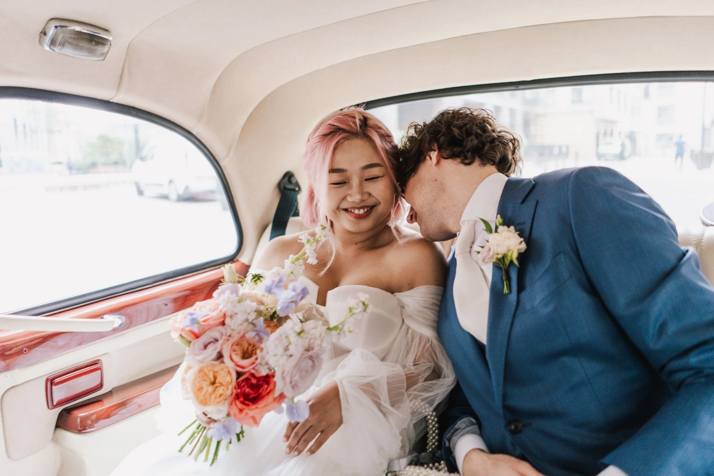 A newly married couple share a tender moment in the back of a classic wedding car, the groom leaning in affectionately as the bride smiles softly, bouquet resting in her lap. Her pastel-coloured flowers and romantic off-the-shoulder gown bring a drea