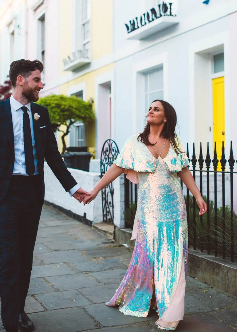 A stylish and contemporary London wedding portrait captured by Barbara K Photography, featuring a couple walking hand-in-hand down a pastel-lined residential street. The bride wears a dazzling iridescent sequin gown that catches the evening light bea