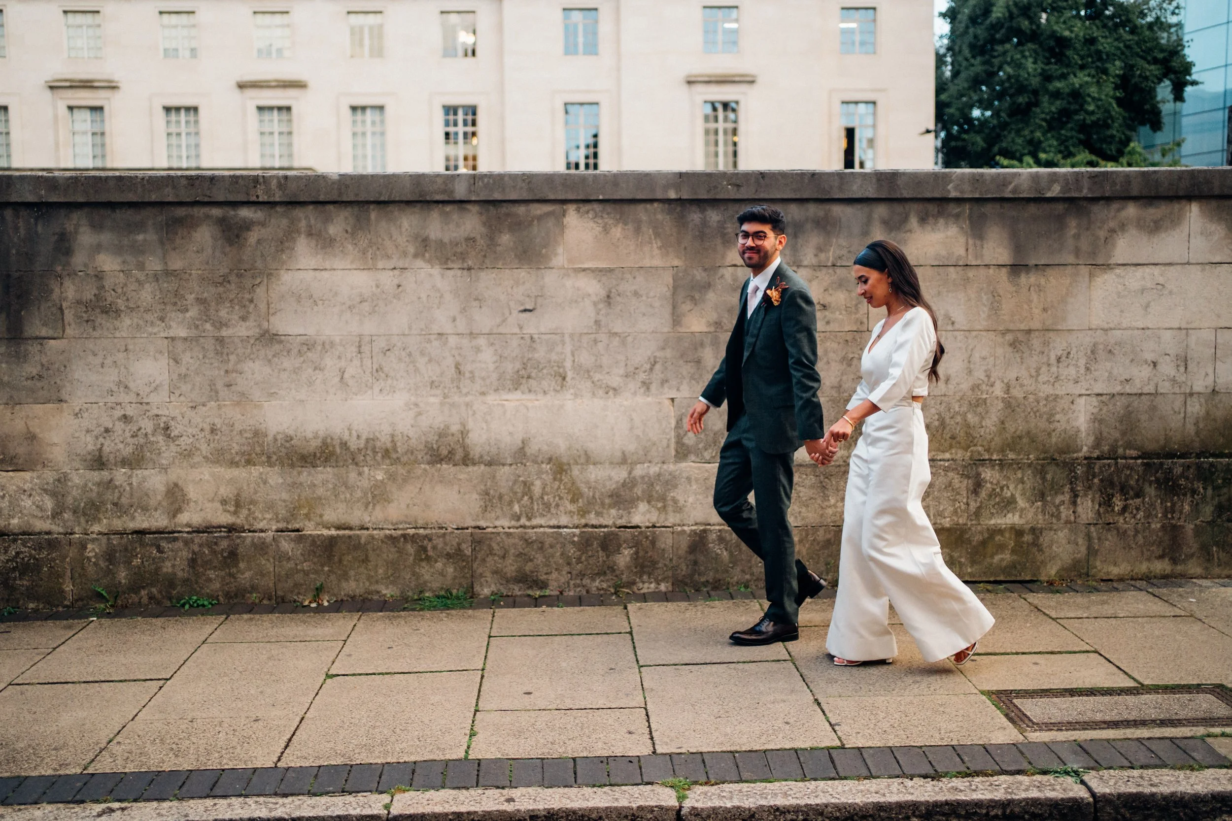 The image shows a couple walking hand in hand along a paved city street beside a tall stone wall, with the façade of a large building and some greenery visible in the background. The woman is wearing an ivory bridal two piece outfit with a V neck top