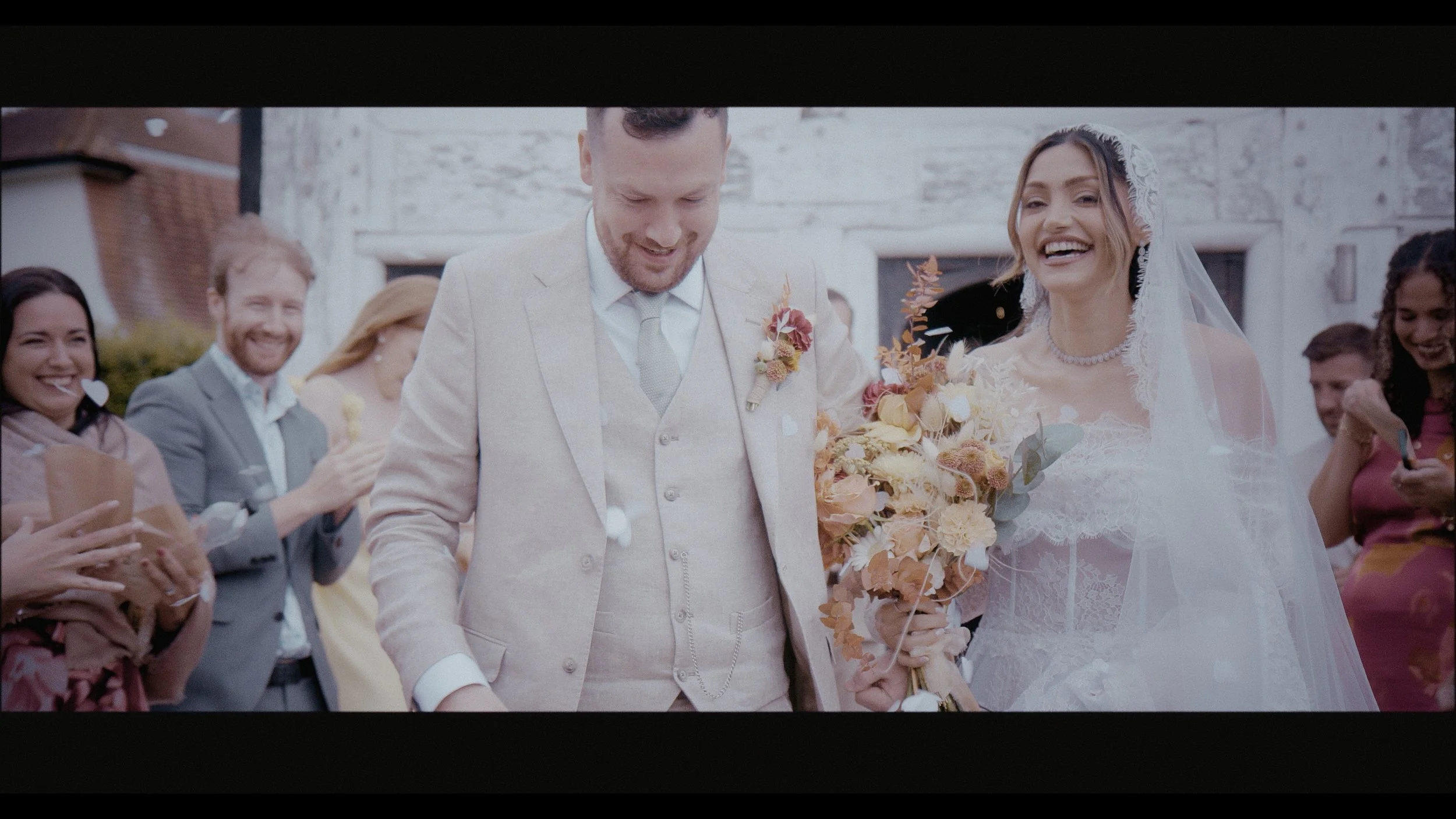 A joyful newlywed moment filmed by Heart Shaped Video, capturing the couple walking through a flurry of confetti with laughter and love. The groom’s soft beige suit and the bride’s lace gown and veil perfectly complement the warm autumnal florals in 