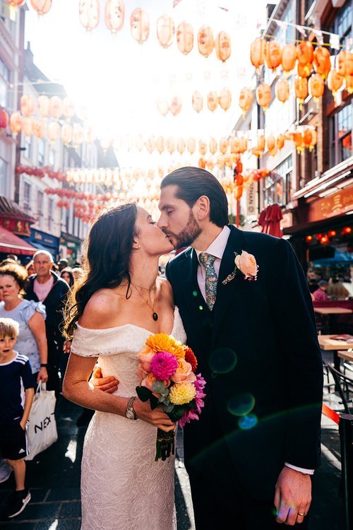 A newly married couple share a kiss in a vibrant city street lined with hanging lanterns, beautifully captured by Jordanna Marston Photography. The warm sunlight, colourful surroundings and lively atmosphere create an energetic and joyful backdrop th