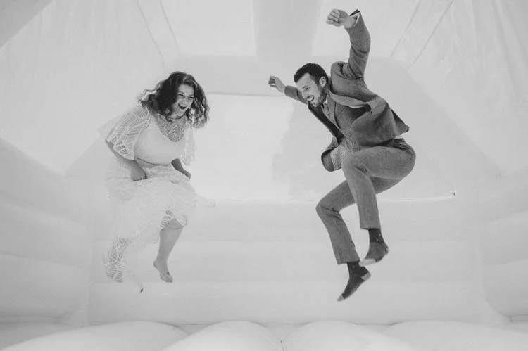 A fun and carefree wedding moment captured by Matt Fox Photography, showing a couple joyfully bouncing together on a white inflatable castle. The bride’s flowing dress and the groom’s suit create a playful contrast as they lift off the ground, both l
