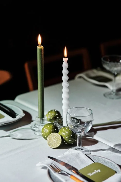 A stylish and moody wedding tablescape styled by Orange Tree Weddings, featuring green and white twisted candles, fresh limes, and minimal glassware on crisp white linens. The modern citrus-inspired design adds an elegant and contemporary feel to cre