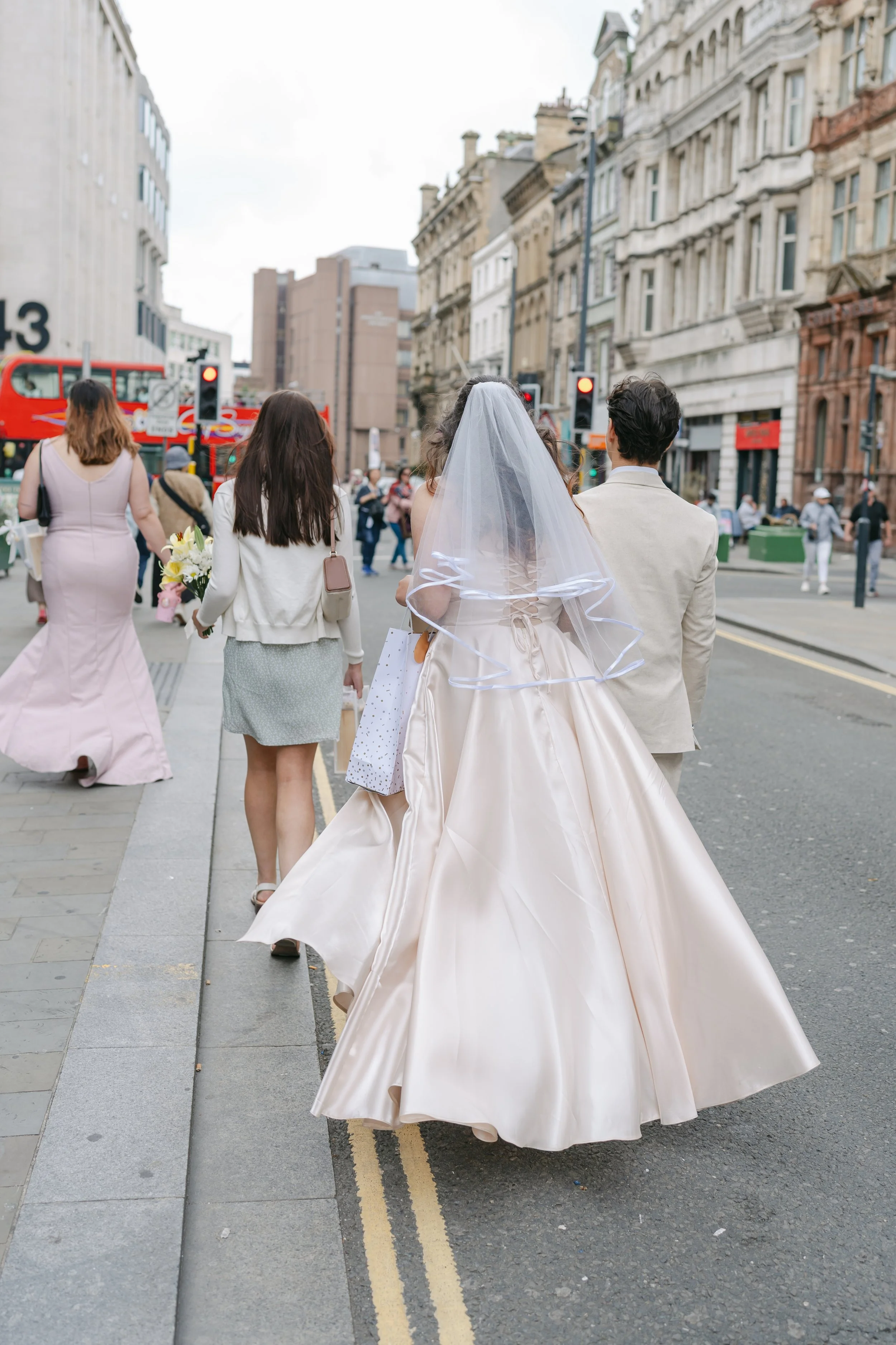 A bride walks through a busy city street with her wedding party, her satin gown and veil catching the movement of the day, beautifully captured by Christy Photography. Surrounded by historic architecture, red buses and the energy of urban life, the s