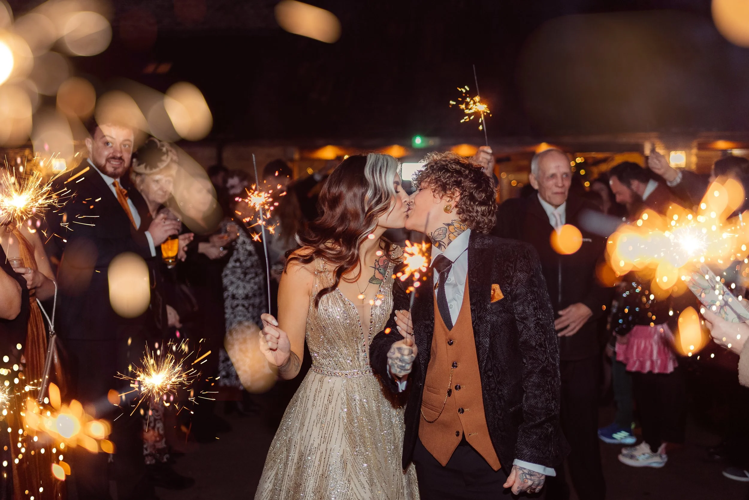 A vibrant sparkler exit captured by Gemma Gaskins Photography showing a newly married couple sharing a kiss surrounded by guests holding glowing sparklers. The warm lights, joyful crowd and candid documentary style create a magical and energetic cele