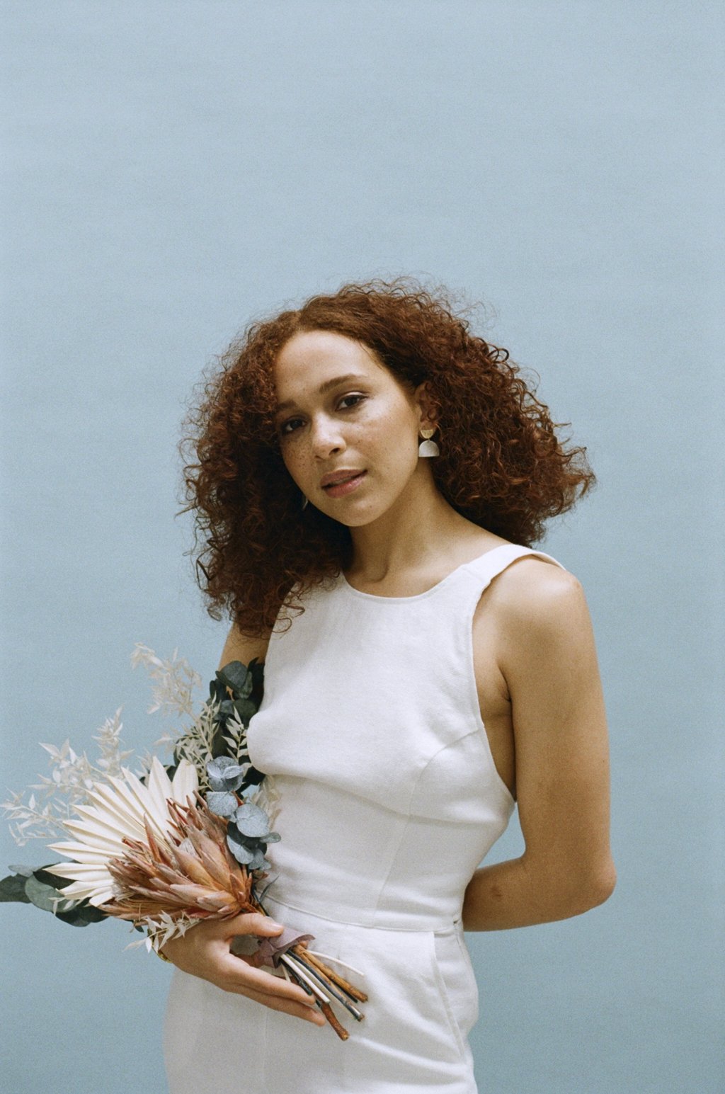 The image shows a woman standing against a soft blue backdrop wearing an ivory sleeveless bridal jumpsuit with a fitted bodice and a high round neckline while holding a small bouquet of dried and fresh botanical stems including protea flowers and euc