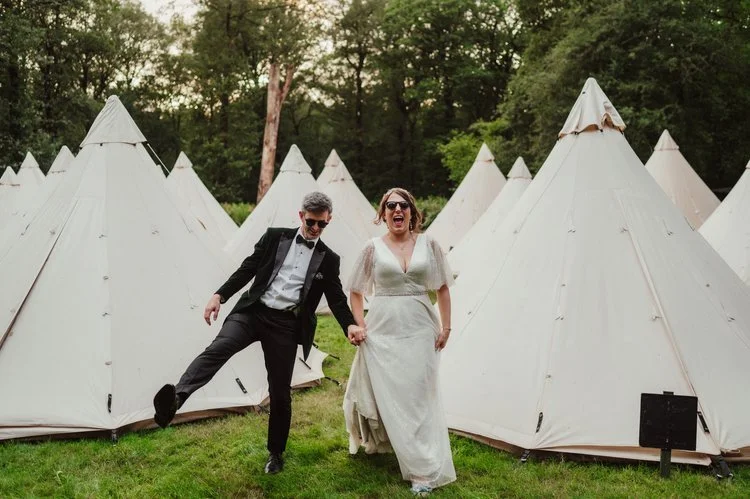 A joyful couple walk hand in hand through a field of cream-coloured bell tents at their outdoor festival-style wedding, laughing together as they move across the grass. The bride’s relaxed, flowing gown and the groom’s classic tuxedo create a fun con