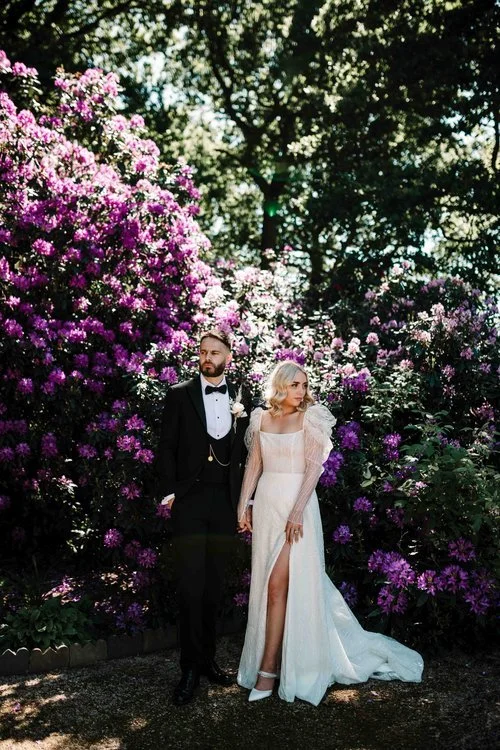 A stylish couple stands framed by dramatic purple rhododendrons, creating a cinematic and fashion-forward wedding portrait. The bride’s ethereal gown, complete with sheer puff sleeves and a thigh-high slit, catches the light beautifully as she holds 