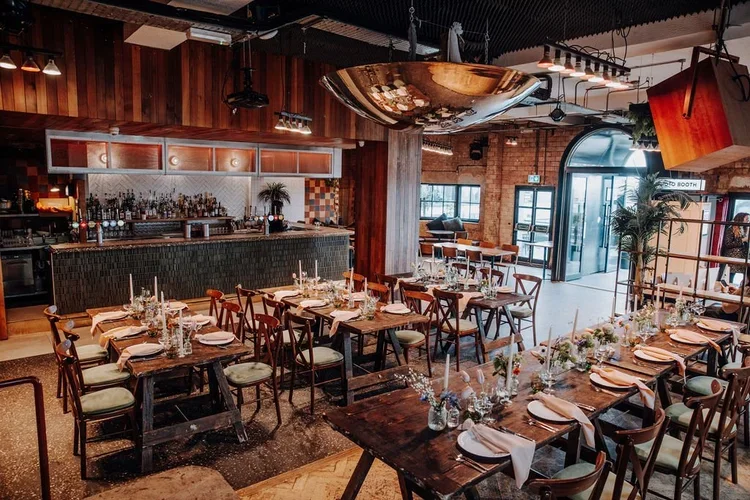 A modern wedding reception setup at Pier Court Brighton, featuring rustic wooden tables, elegant candlelit decor, and an industrial-chic interior with exposed brick and a stylish cocktail bar. Natural light floods the space, creating a relaxed coasta