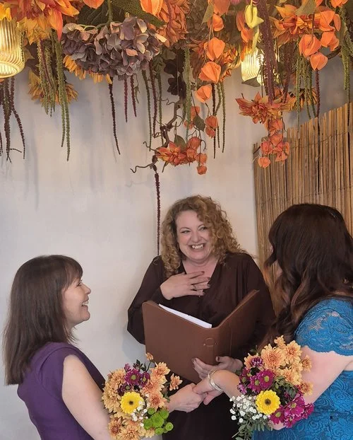 Melly the Celebrant smiling warmly while officiating a ceremony between two brides holding colorful floral bouquets. The setting features a stunning ceiling display of hanging autumnal flowers and soft lighting, creating a romantic, joyful atmosphere