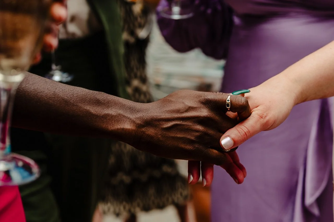 Two hands meet in a tender clasp surrounded by the soft blur of guests and champagne glasses creating a quiet moment of connection that contrasts beautifully with the vibrant colours and movement around them.