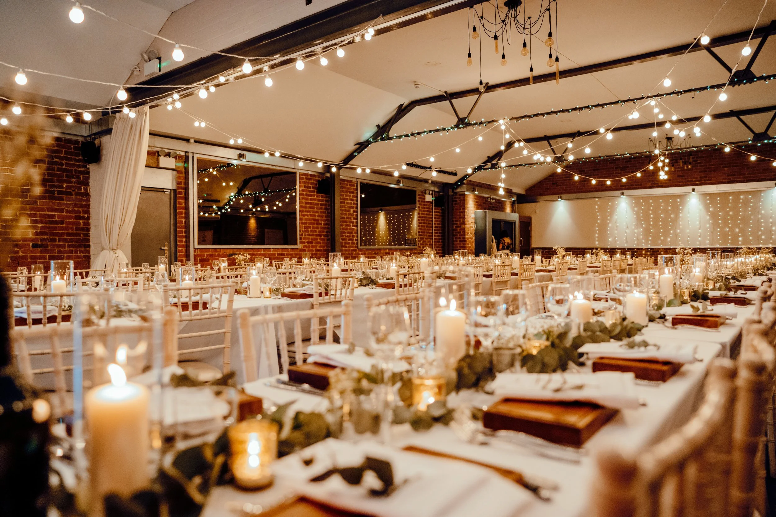 A warm and inviting reception hall is filled with long candlelit tables dressed in white linens, gold cutlery and soft eucalyptus garlands, while rows of light wooden chairs create a sense of symmetry and celebration beneath a ceiling draped with glo