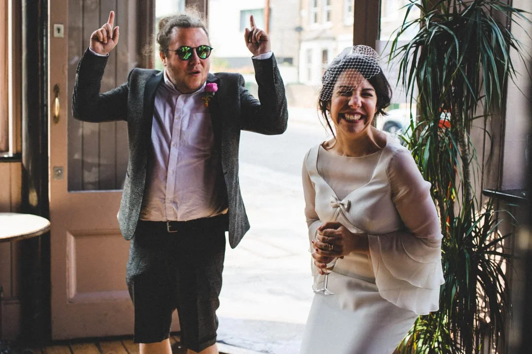 A fun and spontaneous wedding moment captured by Barbara K Photography, showing a couple entering a venue with huge smiles and infectious energy. The bride wears a chic satin dress with sheer sleeves and a vintage inspired birdcage veil, holding a gl