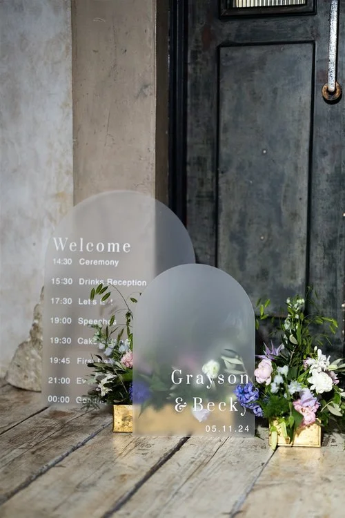 Modern frosted acrylic wedding signage by Blue Pumpkin Calligraphy. Featuring minimalist white lettering and elegant arch shapes, this bespoke welcome sign and order of the day pair beautifully with soft floral arrangements - perfect for contemporary
