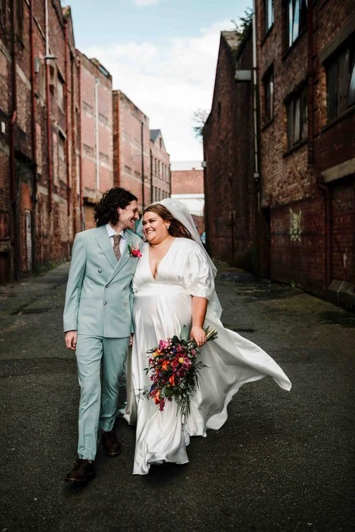 A modern and emotive wedding portrait captured by Amy Faith Photography, showing the couple walking arm-in-arm through a narrow urban alleyway lined with red brick buildings. The groom’s powder blue suit adds a soft pop of colour against the industri