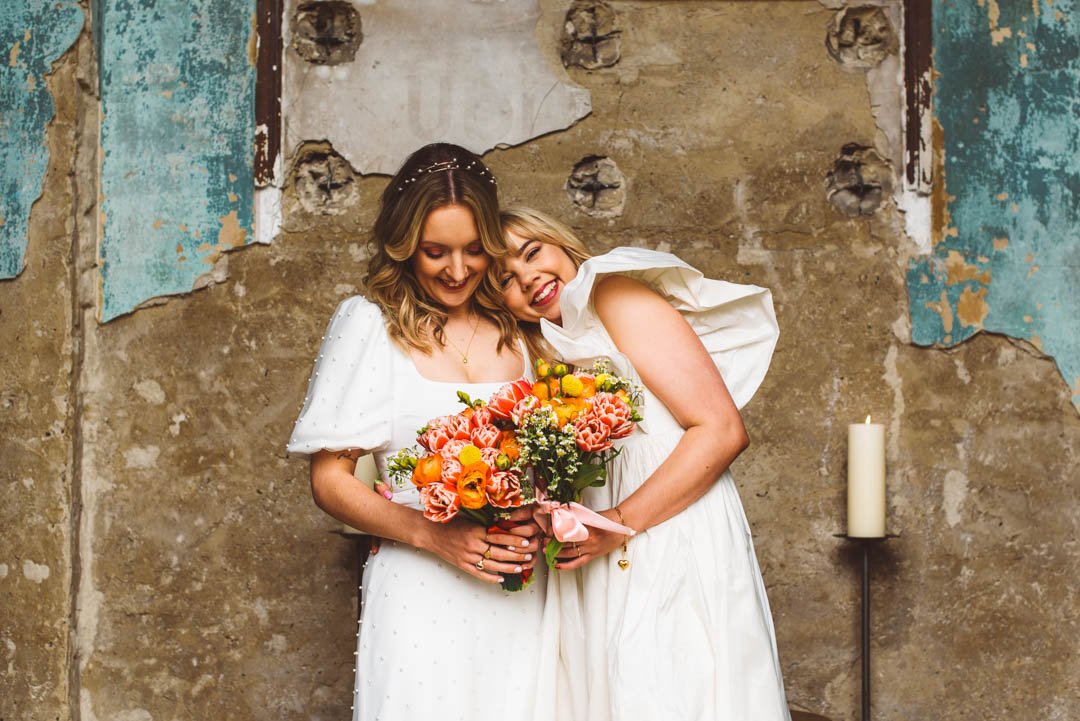 A tender and joy-filled portrait of two brides captured by Barbara K Photography, set against a beautifully weathered, rustic wall that adds character and charm to the moment. Both brides wear elegant modern wedding dresses - one with delicate pearl 